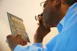 Close-up of a dark-skinned person in a blue shirt examining a small circuit board or electronic component, holding it up close to their face.