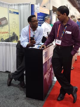 Two African American men in conversation at a conference booth. One seated wearing white shirt, one standing wearing burgundy shirt. Both have name tags. Conference setting with banners and carpet visible.