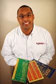 A smiling African-American male with neatly trimmed hair and ๐ wearing business casual clothing including a white button shirt with 'the component store' logo imprinted on it and holding a collection of circuit boards